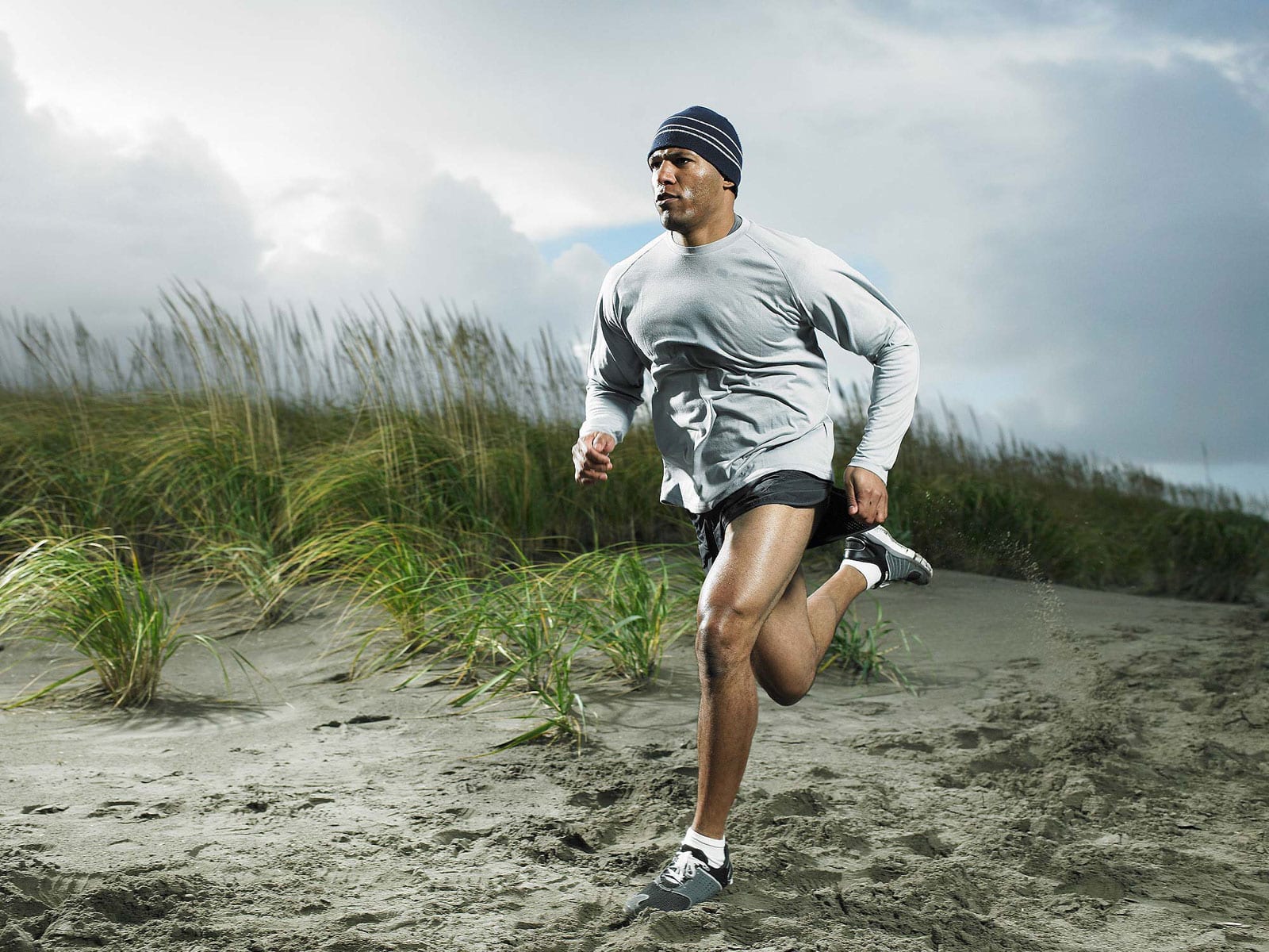 Спортсмен бежит по песку в светлой толстовке и шапке